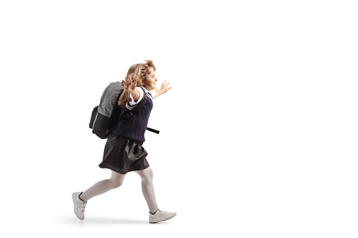 Full length profile shot of a schoolgirl in a uniform carrying a backpack and running with arms wide open isolated on white background