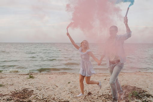Man and woman having fun running on sand and holding colorful blue and red or pink color smoke bombs on the beach. A happy married couple holds hands smoke bomb. Baby shower. Twins:  boy and girl.