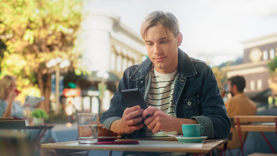 Portrait of a Young Man Typing a Message on a Smartphone Device, Replying to Friends on a Social Network Post. Stylish Male Enjoying a Cup of Coffee at a Cozy City Cafe Terrace