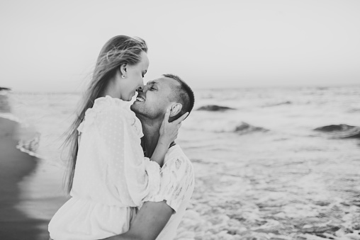 Couple in love hugging and kissing on seashore in sea. Male kisses and hug female standing on water with big waves ocean and enjoying summer day. Man embraces woman on beach sea. Black and white photo