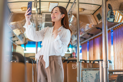 Modern Young Asian Businesswoman, Smart Casual Style, Video Calls and Greets Friend on Phone while Standing on a Public Bus