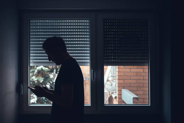 istock Depressed sad man using mobile phone in dark room by the window with shutters pulled down 2150667395