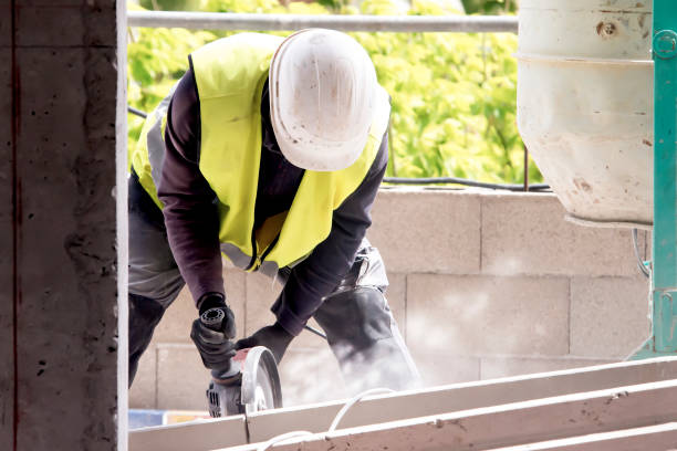 A worker in protective equipment works with a circular machine, cutting the structure, raising dust next to a concrete mixer on a balcony made of concrete blocks of a building under construction