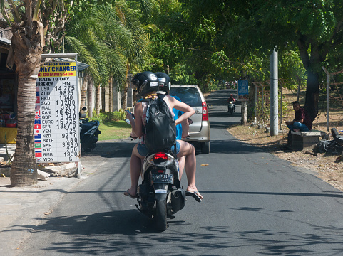 Bali, Indonesia – May 30, 2019: Two tourists on motorbike riding on road with view of international exchange rates on vertical sign.