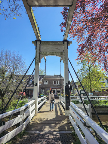 Broek in Waterland Netherlands 21 April 2024, A group of diverse individuals are crossing a majestic bridge over a serene river, each step echoing the rhythm of the flowing water below.