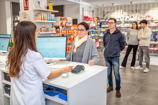 young woman pharmacist helping a people in line while buying medicine at the drugstore - healthcare and medicine concept