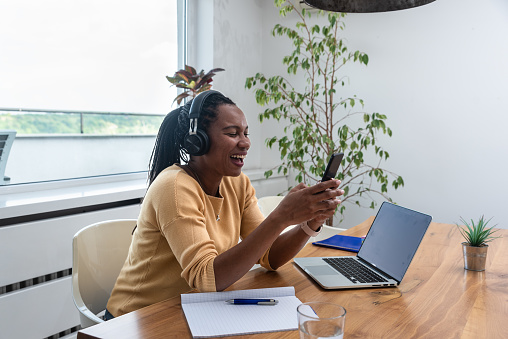 Happy woman wearing headphones sitting with laptop at table in home office. Successful African American freelance business female working online listening the music. Small company owner taking a break
