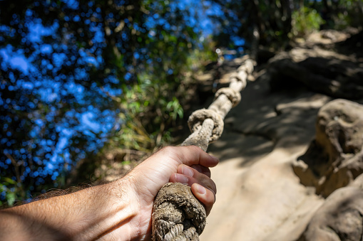 The focus is on a person's hand firmly grasping a weathered rope used for support while climbing a steep trail. The background is a blur of natural elements, indicating a challenging outdoor activity