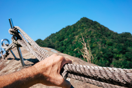 Close up of a male hand reaching for a rope on a via ferrata. In the background is a green overgrown mountain and blue sky as copy space