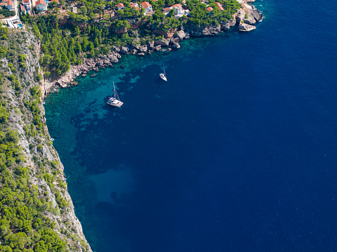 AERIAL: Flying above two sailboats anchored by the coast of Hvar. Anchored yachts rest in the tranquil turquoise waters of a secluded bay, bordered by a rugged cliff and lush Mediterranean greenery.