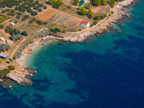 AERIAL: Few camping vehicles and lone house face the turquoise Adriatic sea. Panoramic shot of a remote beach near a camping site on the island of Hvar. Rugged shoreline of a Mediterranean island.