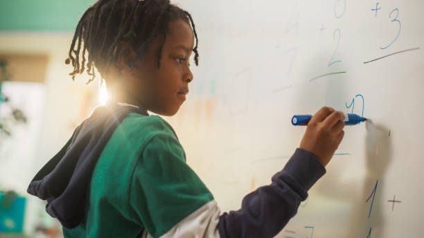 istock Portrait of a Handsome African Boy Finding a Solution to a Mathematical Task on a Whiteboard in Class in Elementary School. Young Pupil is Focused on Numbers, Writing Down a Correct Answer 2150684256