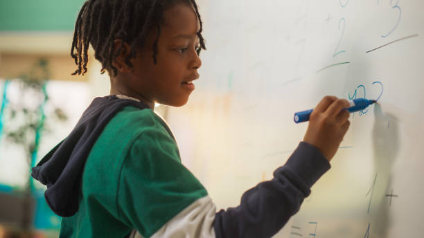 istock Portrait of a Handsome African Boy Finding a Solution to a Mathematical Task on a Whiteboard in Class in Elementary School. Young Pupil is Focused on Numbers, Writing Down a Correct Answer 2150684265