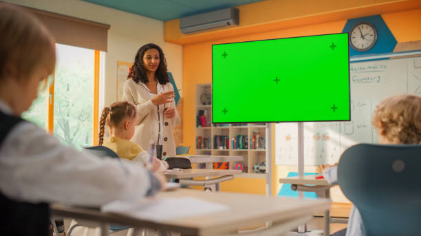 istock Young Teacher Explaining a Class Assignment to Elementary School Kids on a TV Screen with Green Mock Up Chroma Key. Diverse International Kids Receiving Modern Primary Education 2150684431