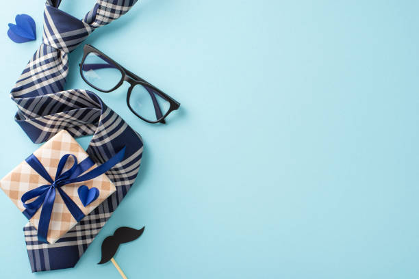 istock Make Dad feel special on Father's Day with heartwarming top view setup featuring classy tie, ribbon-decorated gift box, glasses, mustache props, hearts on pastel blue background, perfect for greetings 2150688870