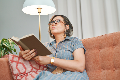 Asian woman, home and bible in living room to relax on day off and leisure. Christian, mature person and religious in  lounge or satisfied on break, chill and self care with positivity and spiritual