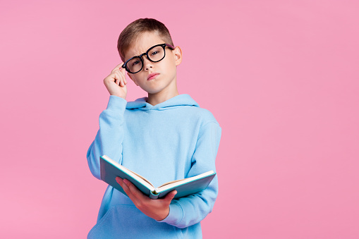 Portrait of small clever minded boy hand touch eyeglasses hold opened book empty space isolated on pink color background.