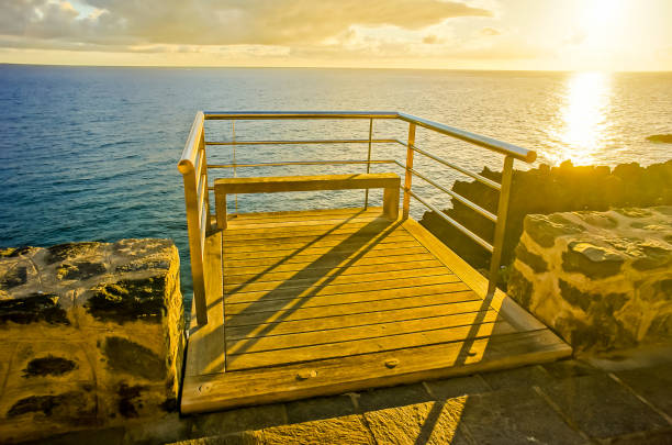 A wooden bench is on a pier overlooking the ocean. The bench is empty and the sky is a beautiful orange color