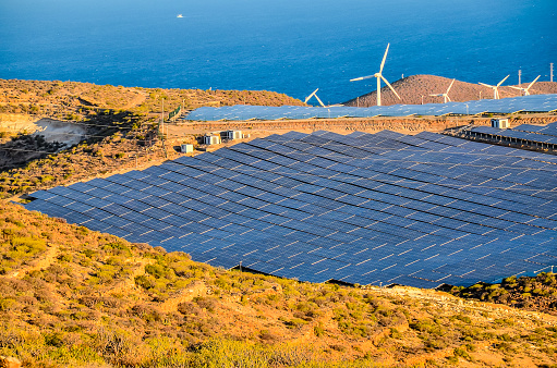 A large solar farm is located on a hillside next to a body of water. The solar panels are spread out across the hill, and there are several wind turbines in the background