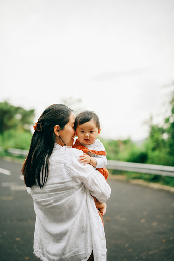 An Asian woman tenderly kisses her smiling child, exuding a warm and loving atmosphere. Surrounded by green trees and a distant hill, the pair stands on a tarred road, enjoying a peaceful moment in a slightly cloudy but bright day.