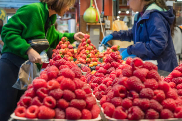 August 10, 2023-Vancouver, BC, Canada-Close up of boxes of colorful fresh organic berries on display at a farmers market with a woman purchasing some items from a fruit seller in background.