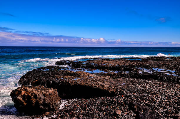 The ocean is calm and the sky is blue. The rocks on the shore are large and the water is clear