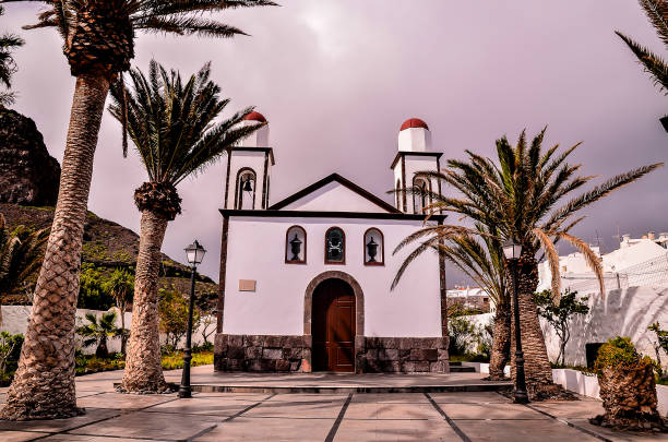 A white church with two red domes sits in front of palm trees. The church is surrounded by a courtyard with a few trees and a bench. The scene has a peaceful and serene atmosphere