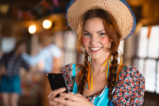 Radiant woman in festive attire interacting with smartphone at Junina celebration. Enthusiastic participant in traditional dress browsing on her phone amidst Brazilian cultural event