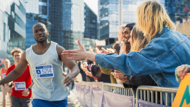 istock Marathon Audience Clapping and Cheering Their Loved Ones Participating in the Race: Black Athletic Male Marathon Runner Giving a High Five to Female Friend Supporting Him in the Audience While Running 2150702746