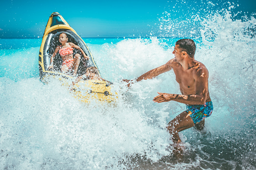 Happy family is enjoying floating in yellow kayak at tropical ocean water during summer vacation. The father is guarding children who are driving in a rubber boat and waves are splashing them.