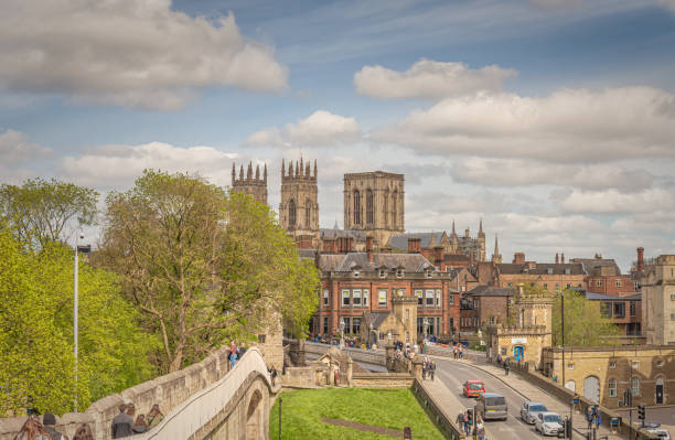 York, UK.  April 20, 2024.  A view of the towers of York Minster rising above the rooftops of the city from the historic city walls.  Tourists walk on the wall and people and traffic cross the bridge.