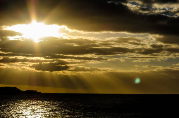 The sky is cloudy and the sun is shining through the clouds. The water is calm and the rocks are scattered along the shore
