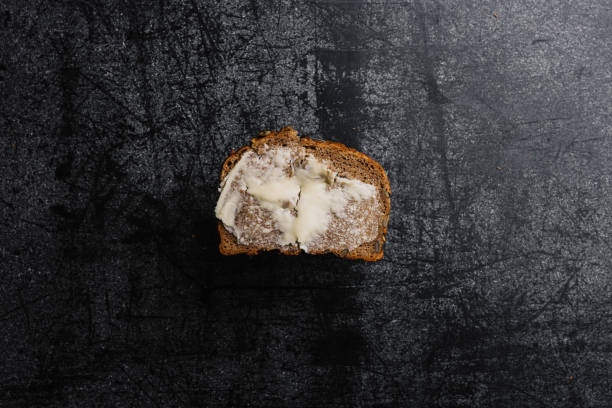 Square slice of fresh bread spread with butter on a dark stone background. The slice of bread lays with the buttered side on top. It is a light farmhouse loaf with a bit of flour on the crust. Photographed from the bird's-eye perspective. Square slice buttered bread still life overhead Studio shot.