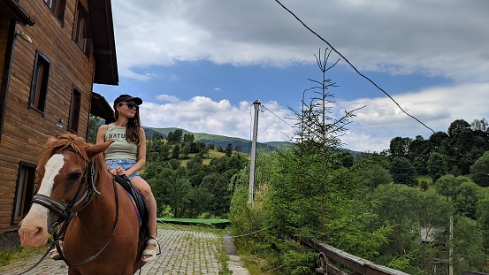 A  girl rides a horse against the backdrop of wooden cottages and a green mountain peak.