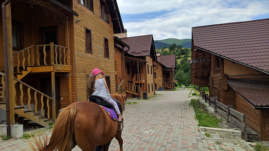 A 5-year-old girl rides a horse against the backdrop of wooden cottages and a green mountain peak.