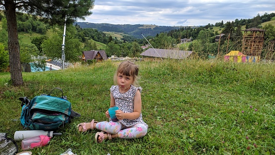 A 5-year-old girl drinks tea near wooden cottages against the backdrop of green mountain slopes and forests.