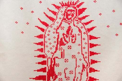A red and white embroidered image of a woman with a crown and a cross. The image is of a religious figure, possibly Mary, and is surrounded by a white background
