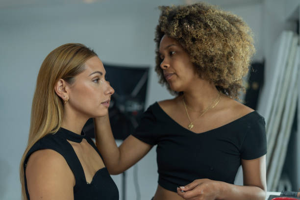 two Latin women dressed casually stand together while one of them with afro hair, a professional makeup artist, attends to her client who attends her beauty business in search of a beautiful social makeup