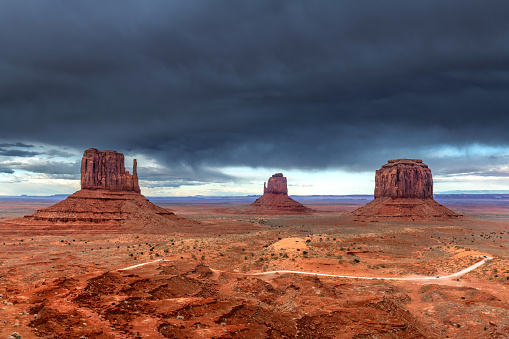 Storm clouds form over East, West and Merrick Butte at Monument Valley while sunshine still penetrates to highlight the vibrant red terrain and roadway.