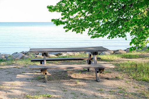 A wooden picnic table sits on the beach next to the water. The table is empty and the grass is green. The scene is peaceful and relaxing, with the sound of the waves in the background.