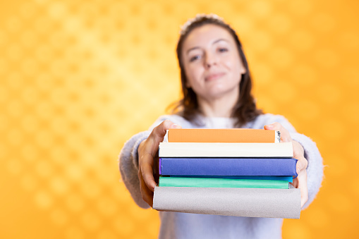 Portrait of smiling woman offering pile of books, recommending reading hobby for relaxation purposes. Radiant bookaholic person with stack of novels in arms doing endorsement, studio background