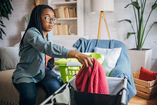 A black woman sits on the sofa of her cozy living room, sorting laundry in a laundry basket.
