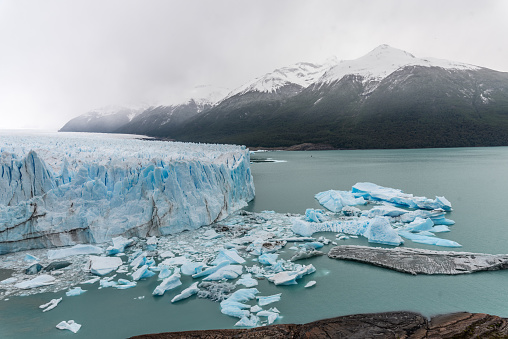 A large body of glacier Perito Moreno is surrounded by icebergs and snow. The sky is cloudy and the mountains in the background are covered in snow. The scene is serene and peaceful, with the ice