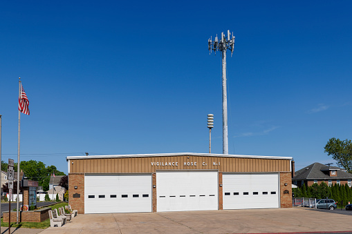 Nazareth, Pennsylvania, USA — May, 26, 2024: Vigilance Hose Co. No.1, a fire station building with a modern design. The building, featuring a beige facade with four closed garage doors, has an American flag waving beside it. A cell tower rises in the background