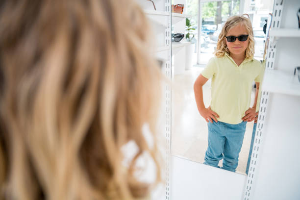Young Caucasian boy with long blonde hair wearing sunglasses stands with hands on hips, reflected in a full-length mirror at an eyewear store.