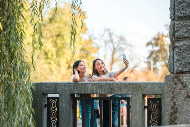 3/4 length image of two attractive Asian female friends taking a selfie in the middle of an old bridge with willow trees hanging over it, both wearing casual clothes  and smiling.