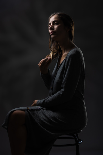 An atmospheric studio portrait showcasing a young woman dressed in a grey dress, sitting thoughtfully with subtle lighting.