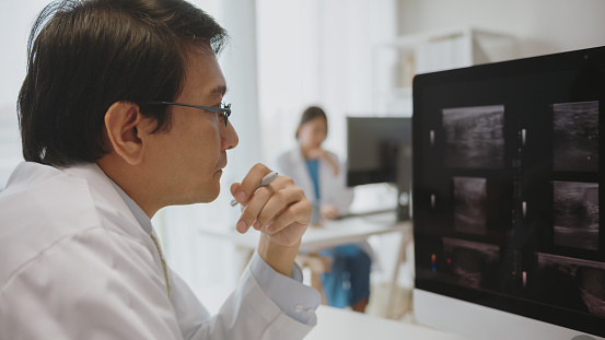 A doctor analyzes medical imaging on a computer, focused intently while consulting with a colleague in the background in a clinic.