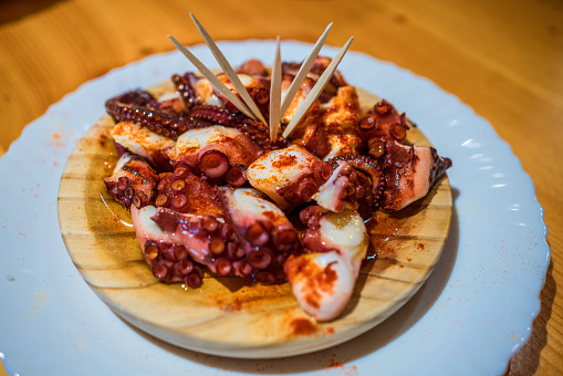 Cooked octopus tentacles seasoned with hot paprika are presented on a rustic wooden plate, each piece skewered with a toothpick, ready for sharing. The dish is arranged enticingly on a wooden table, suggesting a warm, intimate dining atmosphere.