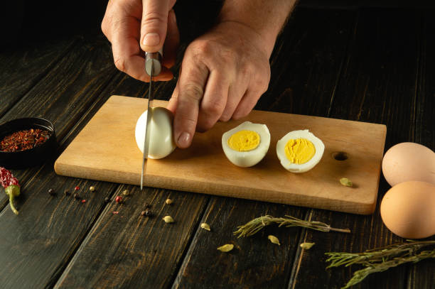 istock Hands with a knife cut an egg. Sliced eggs on a kitchen board for preparing a diet lunch with aromatic spices. Advertising space 2150786263